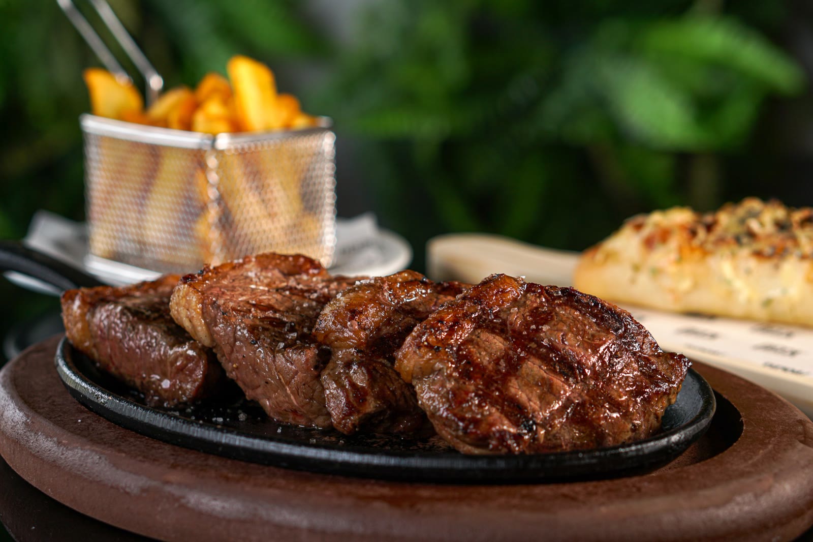 Bife Ancho fatiado na chapa de ferro, com fritas e pão de alho ao fundo