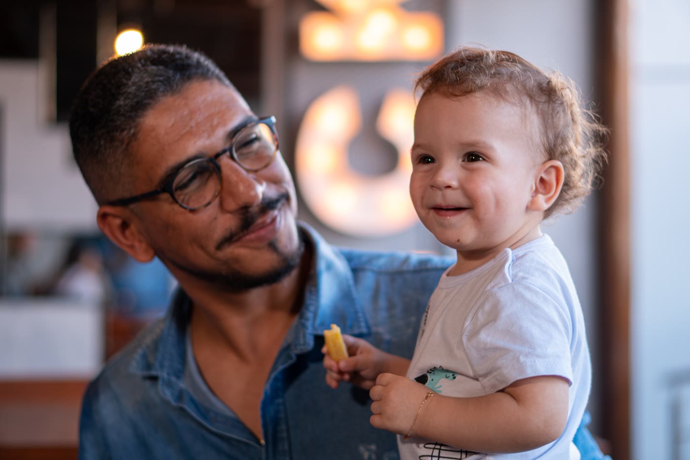 Pai sorrindo para o filho pequeno, que segura uma batata frita
